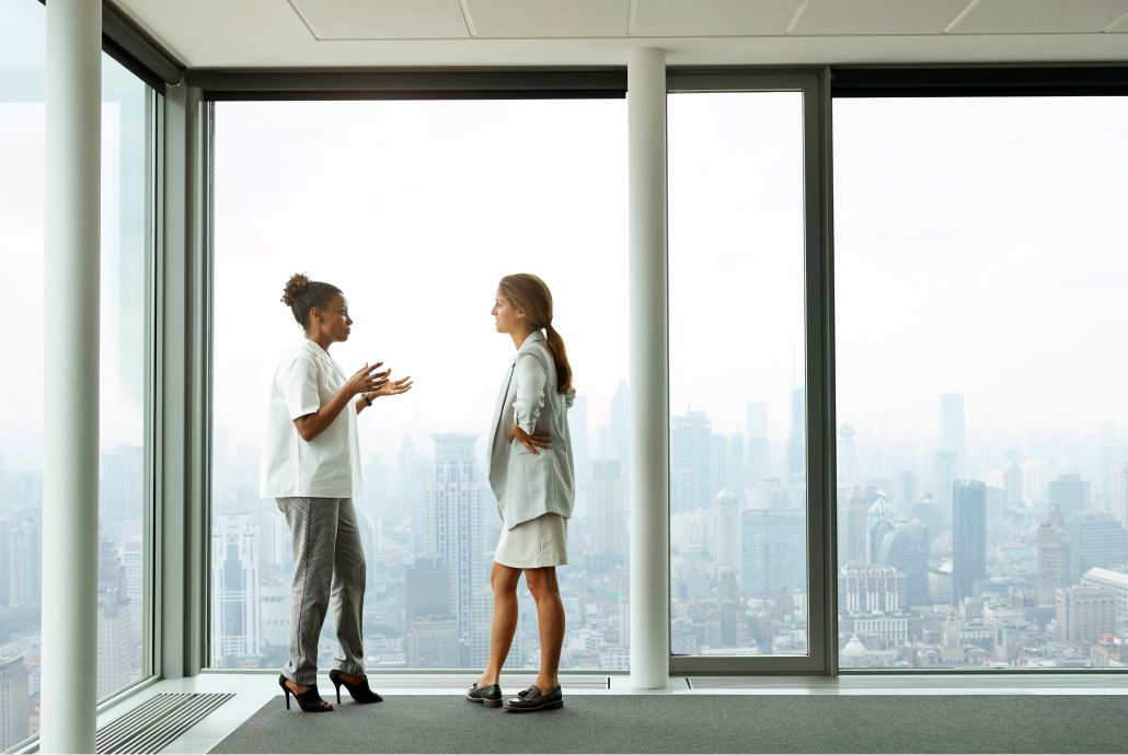 Two women converse in front of a large window showing a city skyline